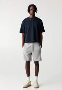 Marineblaues Baumwoll-T-Shirt mit kurzen Ärmeln und grauen Sweatshorts mit Kordelzug. Enthält weiße Knöchelsocken und beigefarbene Sneakers mit gelben Akzenten.