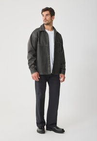 Jachetă din denim neagră cu cusături contrastante, închidere cu nasturi și buzunar pe piept, purtată peste un tricou alb, asociată cu pantaloni închiși la culoare și pantofi.
