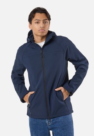 Nuori mies pukeutuneena tummansiniseen hupulliseen softshell-takkiin, jossa on vetoketjulliset taskut, ja sinisiin farkkuihin, seisomassa käsineen taskuissa.