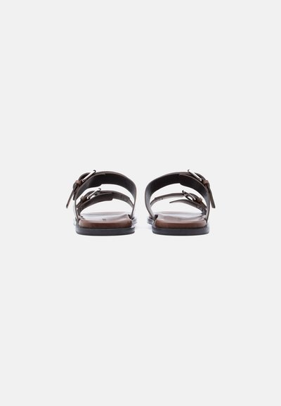 Ζευγάρι καφέ δερμάτινα σανδάλια με επίπεδη σόλα και διπλά λουριά με αγκράφες, εμφανιζόμενα από πίσω σε λευκό φόντο.