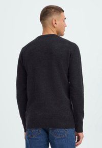 Man met kort haar in een eenvoudige zwarte trui en blauwe spijkerbroek, van achteren gezien tegen een effen lichte achtergrond.