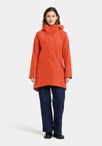 Manteau imperméable orange avec un col haut et une capuche, doté de poches avant et d'un ourlet arrière plus long, associé à un jean bleu à jambes larges.