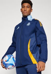 Mornarska atletska jakna sa svijetloplavim detaljima i žutim bočnim detaljima, sa punim zatvaranjem, uspravnim ovratnikom i timskim logotipom na prsima.