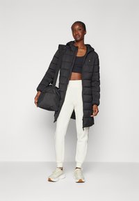 Doudoune noire avec capuche, texture matelassée, fermeture éclair, portée sur un crop top noir et un jogging blanc, complétée par un sac de sport sombre.