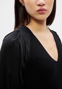 Jauna sieviete ar vidēju ādas toni un taisniem brūniem matiem, valkājot melnu V-veida kakla krekliņu un redzamu ziedu tetovējumu uz krūtīm baltā fonā.