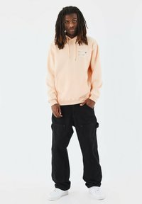Sudadera melocotón con bolsillo delantero y detalle gráfico, combinada con pantalones de cargo negros que tienen bolsillos y zapatillas blancas. Colores sólidos, corte casual.
