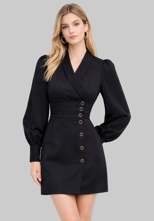 Donna che indossa un mini abito nero a maniche lunghe con colletto incrociato e sei bottoni dorati sul davanti, in piedi contro uno sfondo grigio uniforme.