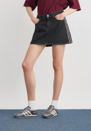 Personne portant une mini-jupe en jean noire avec des bandes latérales, un haut bordeaux, des chaussettes blanches et des baskets Adidas grises, debout sur une moquette bleue.