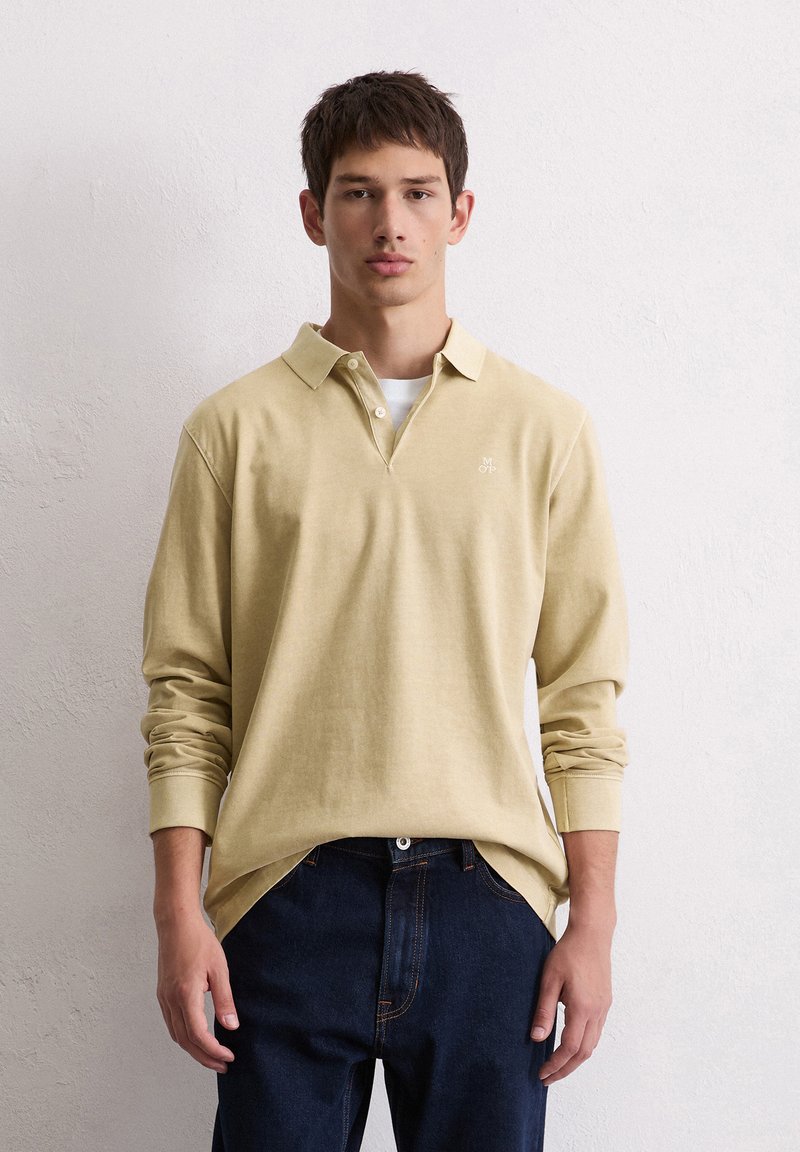 Jasnożółta koszula polo z długim rękawem i kołnierzykiem, z małym logo na piersi. W połączeniu z ciemnymi dżinsami.