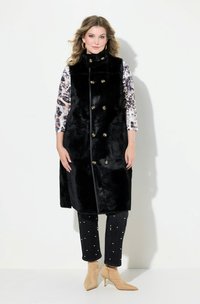 Gilet in pelliccia sintetica nera con colletto alto e bottoni dorati a doppio petto. Indossato sopra una camicia a maniche lunghe a fantasia e abbinato a jeans scuri.