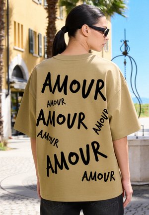 Vrouw met zwarte paardenstaart die een beige oversized overhemd draagt met het woord "AMOUR" meerdere keren zwart gedrukt op de rug.