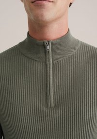Man in een geribbelde olijfgroene trui met een korte rits op de hoge kraag, getoond van schouders tot mond.