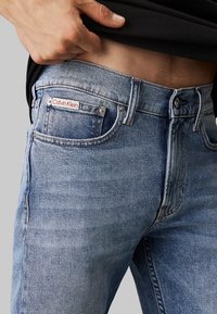 Jasnoniebieskie dżinsy z denimu z czerwonym metką "Calvin Klein". Klasyczny pięcio-kieszeniowy krój z widocznym przeszyciem i wyblakłą teksturą.