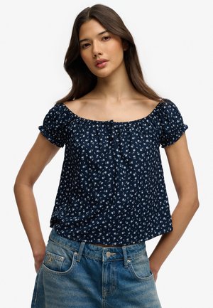 Junge Frau trägt eine marineblaue geblümte schulterfreie Bluse und verwaschene blaue Jeans, steht mit den Händen auf den Hüften.