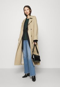 Beige trenchcoat med store knapper, båret over en grøn top og en sort pinstripe blazer. Slidte blå jeans og en sort handbag fuldender looket.
