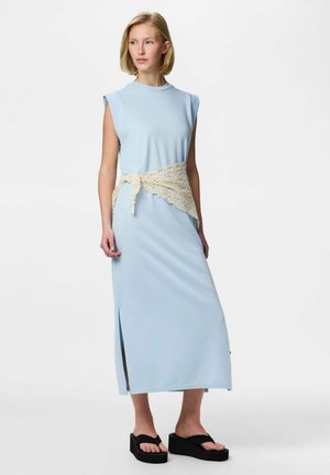 Femme portant une robe midi sans manches bleu clair avec des fentes latérales, une ceinture en crochet crème et des sandales noires à plateforme, debout sur un fond blanc.