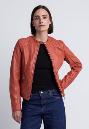 Frau mit kurzen dunklen Haaren, die eine orange Lederjacke mit Reißverschluss, ein schwarzes Oberteil und blaue Jeans trägt, steht mit einer Hand in der Hüfte.