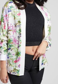 Kvinde iført en sort ærmeløs crop top, blomster mønstret åben skjorte og sorte bukser, holder skjorten åben for at afsløre maven.
