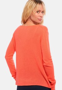 Femme aux cheveux blonds portant un pull orange vif à manches longues, tournée légèrement en arrière, regardant par-dessus son épaule.