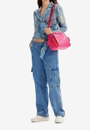Frau trägt eine blaue Wickelbluse mit Muster und lockere blaue Cargo-Jeans und hält eine knallpinke Handtasche mit Schulterriemen.