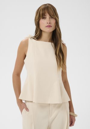 Vrouw met lang bruin haar, gekleed in een mouwloos beige topje met schouderstrikjes en bijpassende beige broek, staand met één hand in de zak.