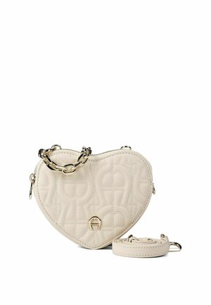 Bolso de mano en forma de corazón color crema con patrón acolchado, asa de cadena tono dorado y correa desmontable a juego colocada a su lado.