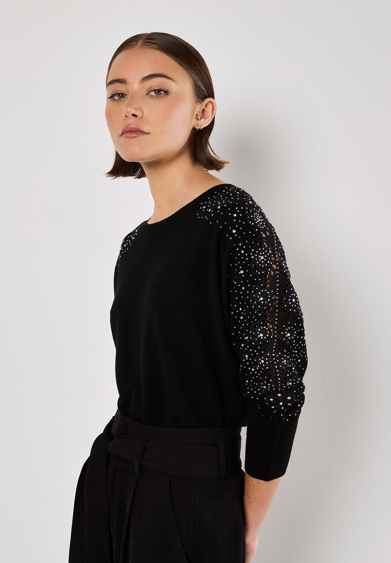 Schwarzer Pullover mit langen Ärmeln, verziert mit funkelnden Strasssteinen an den Schultern, rundem Ausschnitt und weichem Material. Kombiniert mit hochtaillierten Hosen.