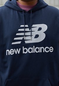 Laivastonsininen huppari, jossa valkoinen "NB"-logo ja "new balance"-teksti, päällä viininpunainen pyöreäkauluksinen paita.