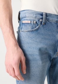 Világoskék farmernadrág egyenes szabással, piros és fehér Calvin Klein logó címkével és látható varrási részletekkel. Strukturált anyag.