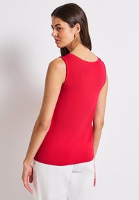 Czerwona bezrękawnik o gładkim materiale, z okrągłym dekoltem i dopasowanym kroju, zestawiony z białymi spodniami.