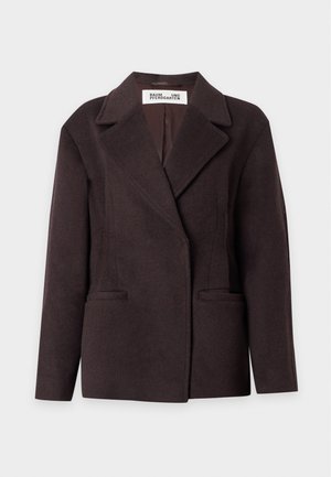 Kvinde iført en mørkebrun oversized blazer, grønstribet sweater, vidde blå jeans og sorte spidse sko, stående med den ene hånd i lommen.