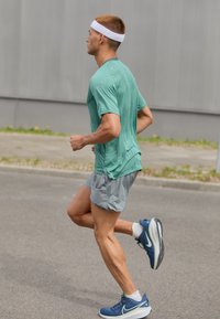 Corridore con una maglietta verde a maniche corte e grigia, che indossa un elastico bianco per capelli e scarpe da ginnastica blu, mentre corre su una superficie asfaltata.