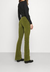 Pantalons évasés en velours côtelé vert avec poches arrière, tissu texturé et détails cousus. Associés à un top noir à manches longues et des bottes.