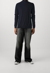 Personne portant un pull à col roulé bleu marine, un jean noir délavé à jambes larges, et des baskets noir et blanc, debout devant un fond clair uni.