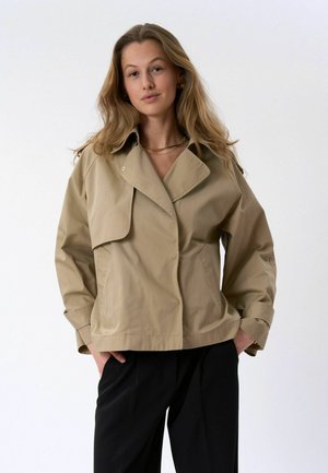 Jeune femme aux cheveux blonds lâchés, portant une veste beige oversized et un pantalon noir, debout les mains dans les poches, fond neutre.