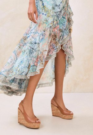 Nainen, jolla on päällä beiget kiilakorkosandaalit ja liehuva korkea-matala kukkakuosinen mekko, seisomassa beigeä teksturoitua taustaa vasten.