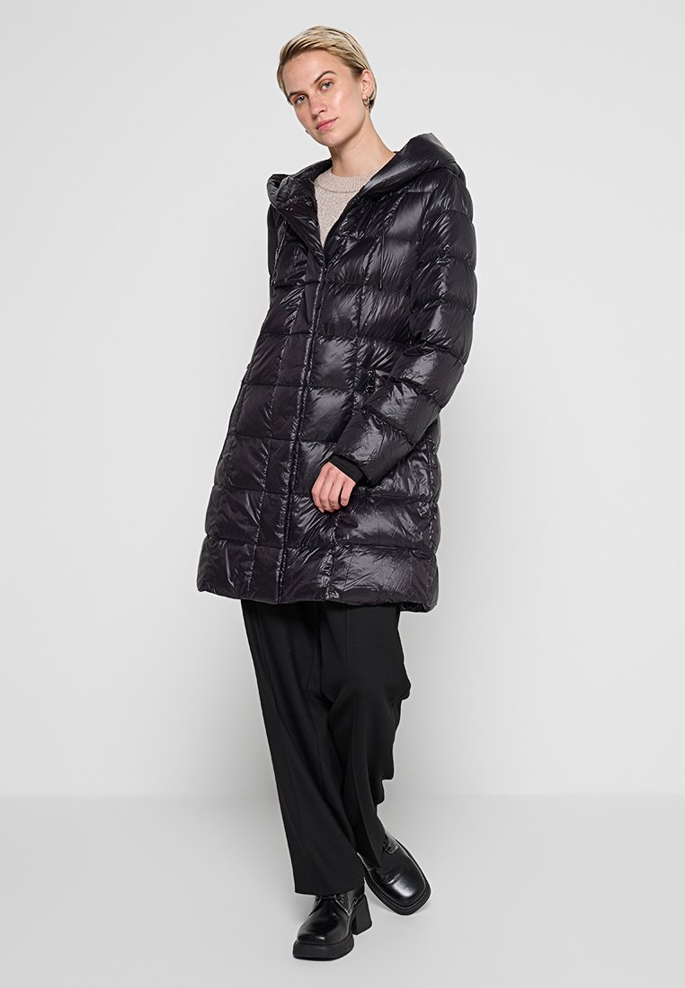 Schwarzer gepolsterter Mantel mit Kapuze, der ein gestepptes Design, glänzende Textur und lange Ärmel aufweist. Getragen über lockeren schwarzen Hosen und Stiefeletten.