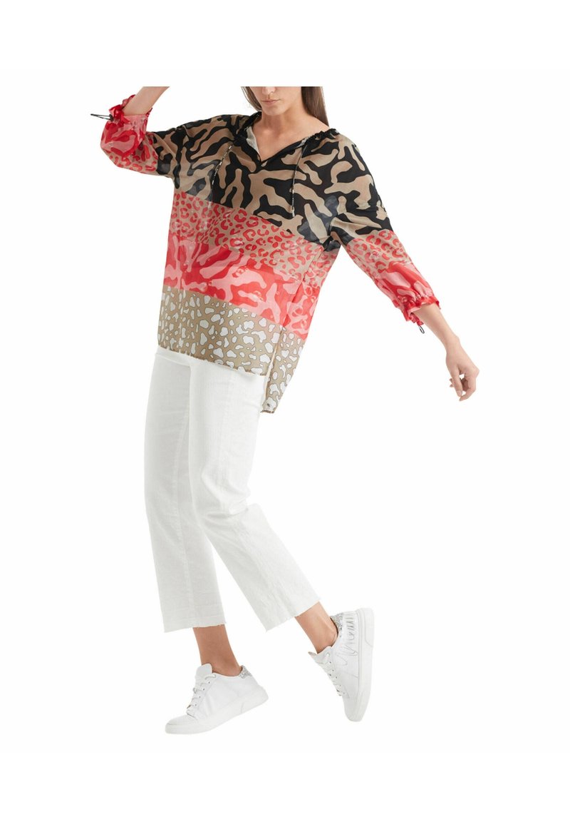 Femme portant un chemisier ample à imprimé animalier multicolore, un pantalon blanc court et des baskets blanches, debout avec une jambe levée sur fond blanc.