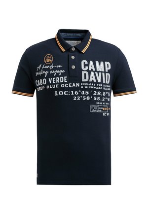 Navy polo shirt with orange striped collar, featuring white printed text and graphics, buttoned placket, and short sleeves. Cotton material.