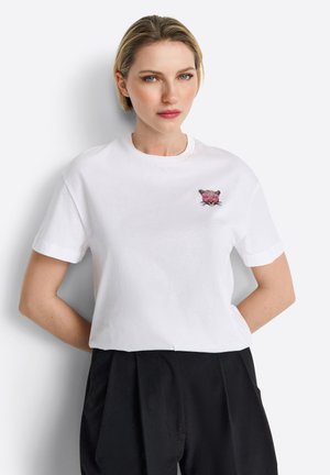 T-shirt en coton blanc avec un motif animal brodé rose et noir sur la poitrine. Manches courtes et coupe col rond classique.