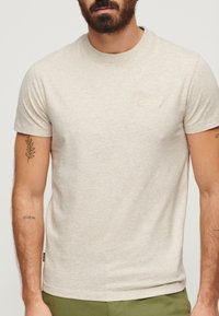 Lys beige t-shirt med korte ærmer lavet af blødt bomuld. Har et diskret mærkelogo på brystet og en ribbet halsudskæring.