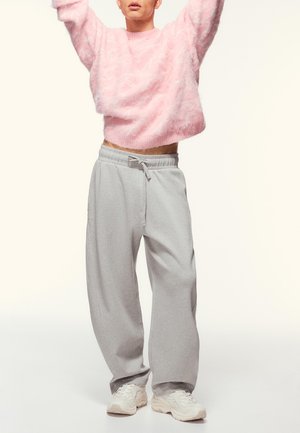 Person, die einen flauschigen rosa Pullover, lockere graue Jogginghosen mit Kordelzug und weiße Turnschuhe trägt, mit erhobenen Armen steht.