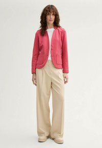 Blazer rose avec deux poches avant, fermeture à un bouton, associé à un pantalon beige à jambes larges et des chaussures chunky beiges.
