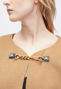 Καστανό ένδυμα από μείγμα μαλλιού με V-λαιμόκοψη, διακοσμημένο με χρυσή αλυσίδα και μαύρες δερμάτινες λεπτομέρειες ασφαλισμένες με κουμπώματα.