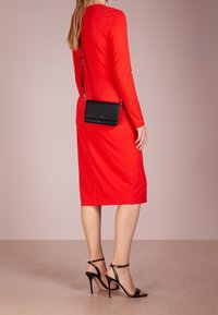 Vestido rojo ajustado con mangas largas, de longitud hasta la rodilla, combinado con tacones negros de tiras y un pequeño bolso cruzado negro. Tejido suave, diseño minimalista.