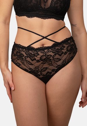 Woman wearing black floral lace high-waist panties with crisscross straps and matching lace bralette, standing against a plain background.