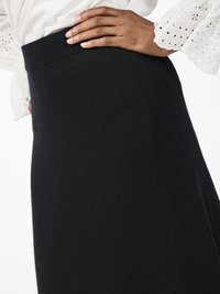 Jupe noire, longueur cheville, avec une taille ajustée et une texture lisse, présentée de côté avec une main reposant sur la hanche.