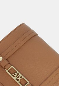 Primer plano de la esquina de una cartera de cuero marrón claro con costuras visibles y un detalle de logo metálico dorado sobre un fondo liso.