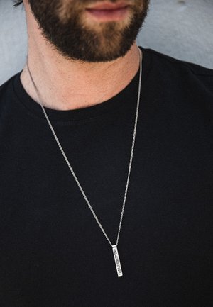 Silverfärgad kedjehalsband med en rektangulär hängande pendant med gravyrtext. Bärs över en svart T-shirt och visar upp en minimalistisk design.