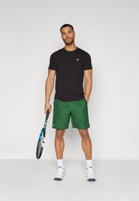 T-shirt noire à manches courtes avec un logo vert, associée à un short vert. Le mannequin tient une raquette de tennis, portant des baskets blanches et des chaussettes.